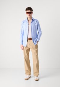 Camisa azul clara de botões, calças bege, tênis brancos com solas em tom de marrom. Acessórios incluem cinto marrom e óculos de sol marrons. Textura suave.