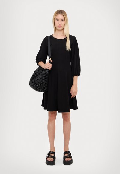 Robe noire à manches courtes avec une jupe évasée, associée à un sac tissé noir à motifs et des sandales noires à semelles épaisses avec des accents de boucle.