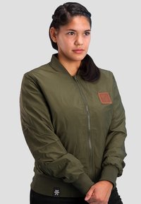 Olive green bomber jacket with a zipper, ribbed cuffs, and a tan leather patch. Smooth texture, lightweight material.