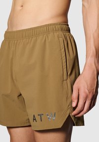 Bruine sportshorts met een elastische tailleband, zijzakken en een reflecterende "ATW" logo-detail op de onderste zoom. Gladde, lichtgewicht stof.