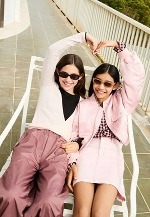 Due ragazze con occhiali da sole, una in una giacca bomber rosa pallido sopra una camicia a quadri bianca e nera, l'altra in un top bianco a coste e pantaloni rosa.