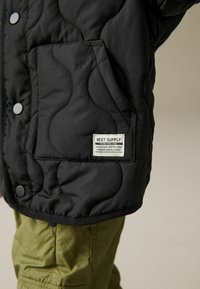 Svart quiltad jacka med en strukturerad yta, med en ficka på bröstet och en etikett som säger "NEXT SUPPLY". Tryckknappar och fållade kanter.