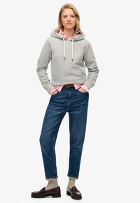 Sudadera gris con capucha, bolsillo frontal, cordones y logotipo; sobre una camisa de color rosa con cuello; combinada con jeans azules y mocasines oscuros.