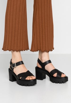 Fødder iført sorte chunky højhælede sandaler med ankelsnører og spænder, sat sammen med brede brune ribstrikkede bukser, der slutter over anklerne.