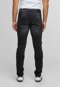 Mees, kes kannab musti kitsaid teksase, valgeid tossusid ja valget ilma varrukateta särki, seisab heleda halli tausta ees.