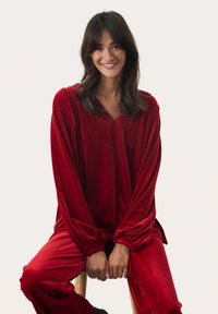 Sada loungewear z červeného sametu obsahující top s V-výstřihem, dlouhými rukávy a elastickými manžetami, doplněná o odpovídající kalhoty s elastickým pasem.