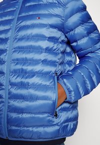 Blaue Steppjacke mit gesteppter Optik, glattem, glänzendem Stoff, Reißverschluss und einem Logodetail auf der Brust. Mit elastischen Bündchen.