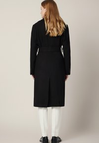 Czarny wełniany płaszcz z paskiem w talii, długimi rękawami, rozcięciem z tyłu i zapięciem na guziki przy mankietach. W połączeniu z białymi spodniami i czarnymi butami.