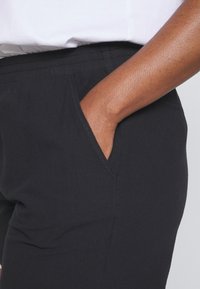 Pantalons de yoga noirs en tissu doux avec une ceinture élastique. Ils sont dotés de poches latérales et d'un design ajusté, partiellement visibles près d'une main de couleur peau.