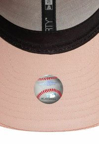 Rosa Baseballmütze mit einem schwarzen Schirm, ausgestattet mit einem runden Etikett, das ein Baseballdesign und Text zeigt. Texturiertes Material mit sichtbaren Nähten.