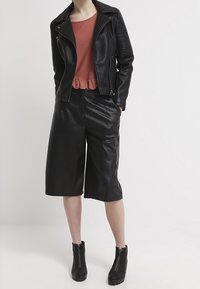 Chaqueta de motorista de piel sintética negra con detalles acanalados en los hombros, combinada con una blusa peplum color óxido y pantalones cortos de piel sintética negra hasta la rodilla. Botines negros.