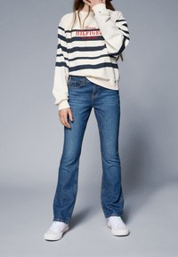 Person klädd i en vit och marinblå randig Tommy Hilfiger sweatshirt, blå jeans och vita sneakers står mot en grå bakgrund.