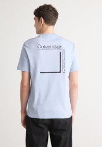 Lyseblå bomulds T-shirt med korte ærmer, med et sort grafisk design og teksten "Calvin Klein" på ryggen, med en rektangulær accent.
