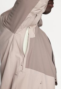 Lightweight, beige jacket with a zippered front, adjustable drawstrings, and contrasting panel design. Smooth, breathable fabric.