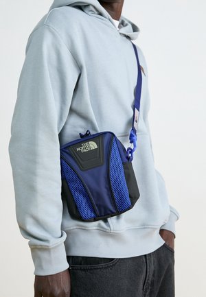 Personne portant un sweat à capuche gris clair et un pantalon sombre, avec un sac bandoulière The North Face bleu et noir à panneaux en mesh, porté sur l'épaule.