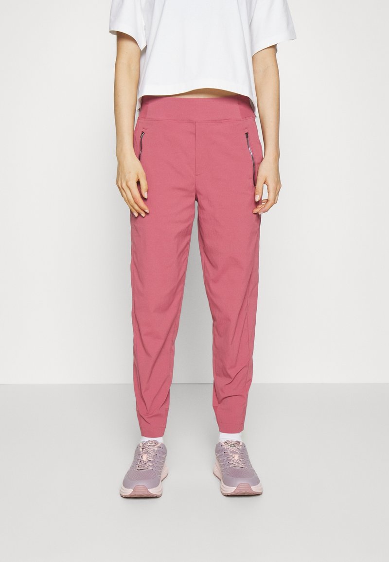 Personne portant un pantalon rose à coupe fuselée avec des poches latérales zippées, un haut blanc court à manches courtes, des chaussettes blanches et des baskets violet clair.