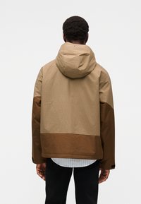 Μπουφάν με κουκούλα σε σκούρο μπεζ και καφέ, με υφή στην πάνω πλευρά, λεία επιφάνεια στο κάτω μέρος και ορατό λογότυπο στον λαιμό. Εξοπλισμένο με ρυθμιζόμενα μανίκια.
