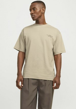 Beige Baumwoll-T-Shirt mit kurzen Ärmeln, rundem Ausschnitt und Siebdruckdetails auf der Brust. Kombiniert mit lockeren grauen Hosen.