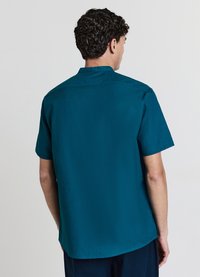 Chemise courte à manches courtes de couleur sarcelle avec un col à revers et une texture lisse. Présente un ourlet droit et un minimum de détails de couture à l'arrière.