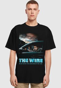 Svart bomull T-skjorte med grafisk trykk av "The Wire," som viser karakterer i en bil med blå tekstlogo og slagord nedenfor.
