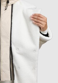 Bílý kabát z fleecu s kulatým límečkem, se dvěma velkými knoflíky a hladkou vnitřní podšívkou, otevřený tak, aby odhalil béžovou izolovanou vrstvu.