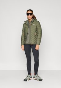 Siltināta olīvzaļu pūkaina jaka ar kapuci, ar oranžiem akcentiem, apvienota ar tumši pelēku kreklu un melnu legingu, piešķirti ar pelēku pārgājienu apavu.