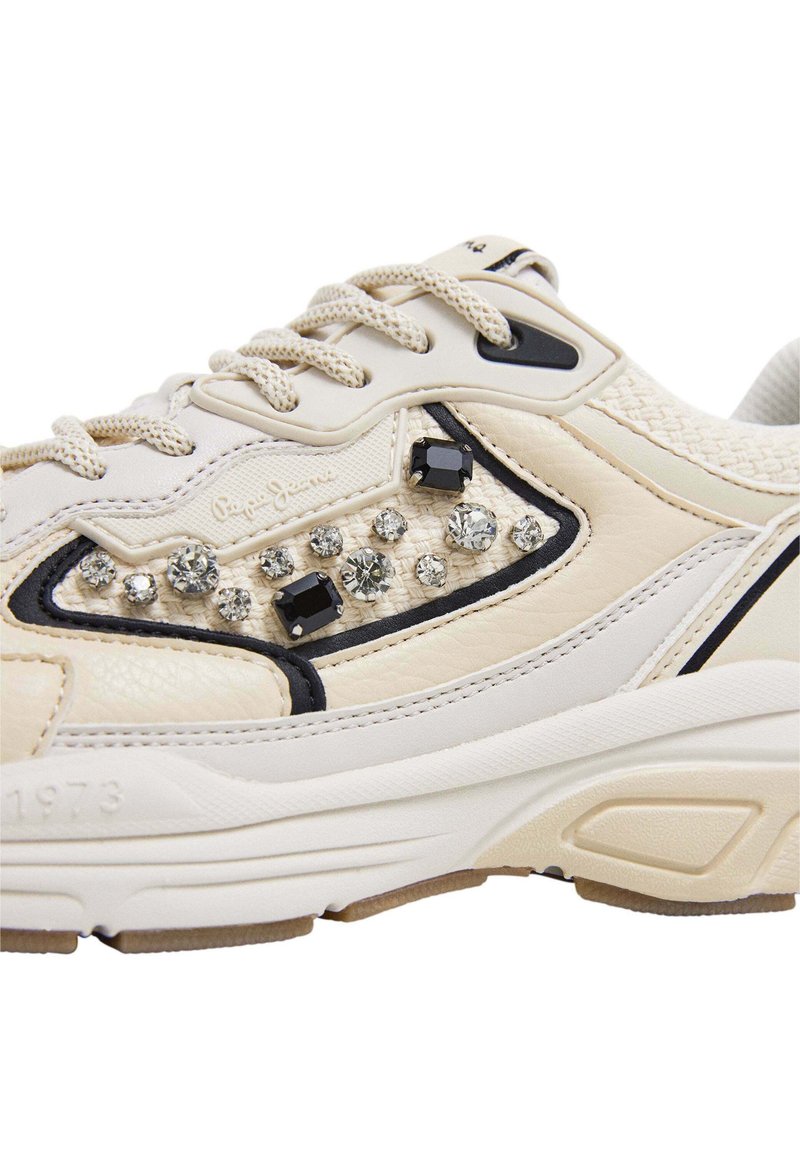 Sneaker beige con tomaia tessurizzata, accenti neri e strass decorativi. Caratterizzato da chiusura con lacci e suola ammortizzata per il massimo comfort.