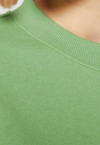 Tela de punto acanalado verde de una camiseta de cuello redondo, que muestra un primer plano del escote y la textura, con un aspecto suave y uniforme.