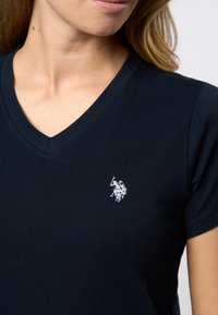Mørkeblå v-hals t-skjorte laget av myk bomull, med en liten hvit brodert logo på venstre bryst.