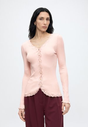 Kvinde iført en lyserød ribbet cardigan med blonderkant og knapper, kombineret med højttaljede mørkerøde bukser, stående mod en ensfarvet baggrund.