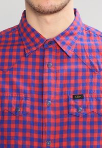 Mann trägt ein rot-blau kariertes Hemd mit Druckknöpfen und zwei Brusttaschen, eine davon mit dem Aufdruck "Lee."