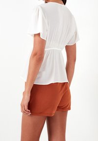 Blusa bianca a maniche corte con vita arricciata e silhouette fluida, abbinata a pantaloni corti color ruggine, vista da dietro.