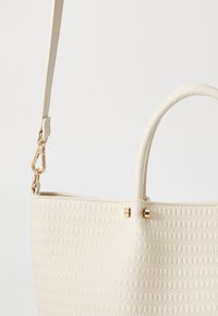 Biała teksturowana torba typu tote z plecionym wzorem, wyposażona w płaski pasek oraz złote akcenty w postaci elementów ozdobnych.