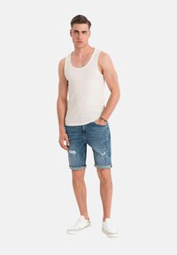 Witte tanktop, denim shorts met opgerolde boorden en versleten details, gecombineerd met witte sneakers. Model staat tegen een lichte achtergrond.