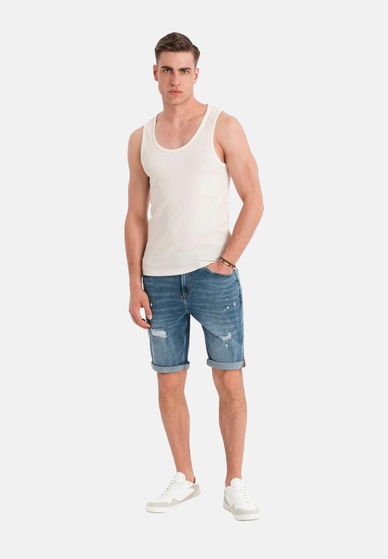 Witte tanktop, denim shorts met opgerolde boorden en versleten details, gecombineerd met witte sneakers. Model staat tegen een lichte achtergrond.
