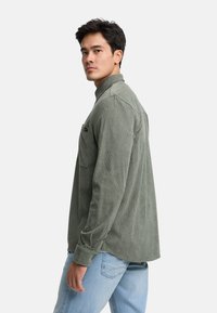 Camisa de pana verde oliva de manga larga con cuello y un bolsillo en el pecho, combinada con unos vaqueros azul claro, vista de lado.