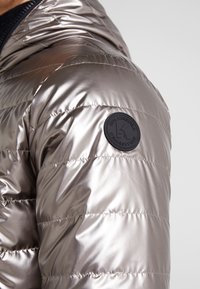 Ärmel einer glänzenden silbernen Steppjacke mit rundem schwarzem Patch-Logo am oberen Arm, mit gesteppten horizontalen Linien und glatter Stoffstruktur.