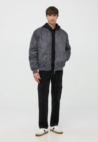 Teksturowana szara kurtka bomberka z prążkowanymi mankietami, nałożona na czarną bluzę z kapturem, zestawiona z czarnymi spodniami cargo i białymi sneakersami.