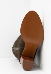 Botte en daim à semelle brune reposant sur le côté contre un mur blanc d’angle, montrant les détails de la semelle et la marque de taille 37.