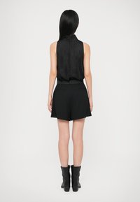 Vrouw met lang zwart haar, gekleed in een mouwloos zwart topje, zwarte hooggetailleerde shorts en zwarte enkellaarzen, staand met haar rug naar de kijker.
