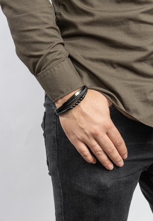 Black leather bracelet with braided texture, silver accents, and round beads. Worn on a wrist, paired with a long-sleeve olive shirt and dark jeans.