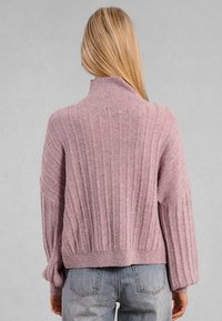 Donna con lunghi capelli biondi, che indossa un maglione a collo alto rigato di un leggero viola e jeans blu, vista da dietro su uno sfondo grigio.