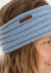 Una fascia per capelli di colore azzurro chiaro presenta un design a coste, realizzata in morbido filato, con un piccolo tag in pelle marrone che riporta il nome del brand.