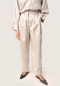 Beige satin vida byxor med hög midja och veck fram. Bärs tillsammans med svarta slingback-skor med spetsig tå.