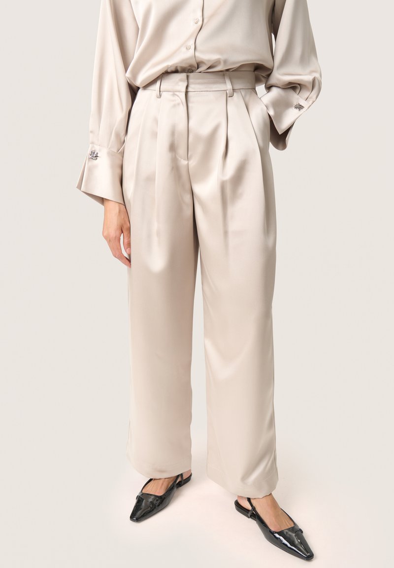 Beige satin vida byxor med hög midja och veck fram. Bärs tillsammans med svarta slingback-skor med spetsig tå.