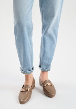 Mocassini in pelle marrone con un accento in catena dorata, abbinati a jeans in denim azzurro chiaro arrotolati. Design casual e piatto con una texture liscia.