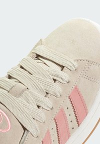 Beige suede sneaker with cream laces, pink zigzag stripes, and perforated accents. Features a textured outsole and a rounded toe shape.