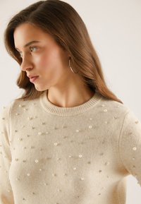 Beige knit sweater featuring a crew neckline, embellished with metallic and pearl-like beads in varying sizes across the front.