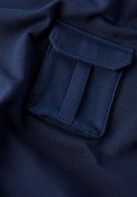 Tissu bleu marine avec une texture lisse, comportant une poche rectangulaire avec des bords cousus et deux fentes verticales pour les objets.