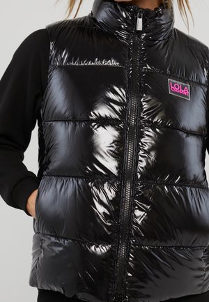 Sort skinnende dunvest med frontlynlås og pink "LOLA LSDMNT" logo-patch, båret over sort langærmet trøje.